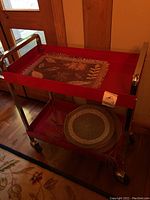 Red metal two-tier rolling bar cart with placemats and bar seat covers placed on it, showing overall condition and size.