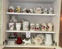 Multiple shelves of assorted mugs, teapots, pitchers, and kitchen items in display cabinet, showing floral patterned porcelain and bone china items.