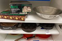 Shelf view showing square baking pan, aebleskiver pan (cast iron with multiple indentations), other cast iron pans, and packaged microwave safe set.