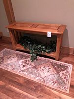 Wooden console table with natural wood grain finish and patterned rug in front on wood floor, includes greenery under table.