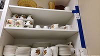 Shelves with stacked plates, cups, and saucers in English Harvest pattern with colorful fruit and gold rim details.