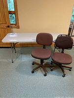 Plastic white folding table and two brown rolling office chairs positioned side by side in front of a beige wall.