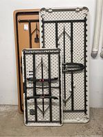 Three folding tables folded and stacked upright against a wall: one wood/particle board 6 ft table, one large white plastic 6 ft table, and one smaller white plastic 4 ft table.