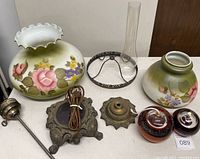 Disassembled gas lamp parts including floral glass shades, metal bases, and glass chimney arranged on a white table.