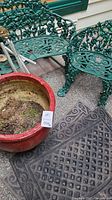 Wide view showing green iron benches with decorative leaf and grapevine design, red flower pot with soil, and two black rubber door mats on concrete floor.