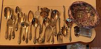 Assorted silver plated serving ware and utensils laid out on a table including spoons, forks, butter knives, and servers