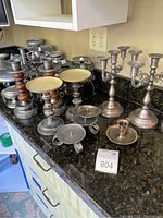 Multiple metal and wood candelabras and candle stands in varying sizes arranged on a black granite counter.