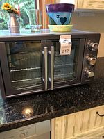 Oster French door convection oven on granite countertop with sunlight reflection. Small vase with sunflowers, glass cup, and colorful ceramic pots on top of oven.