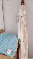 View of closed white fabric patio umbrella with wooden handle next to a stack of blankets including a blue quilted one.