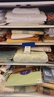Four shelves of assorted folded table linens, mostly white and cream tones with some pastel colors, some in original packaging.