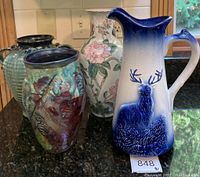 Four assorted decorative vases including a green ribbed vase, a multicolored glazed vase, a floral painted vase, and a blue and white pitcher with deer motif on a kitchen counter.