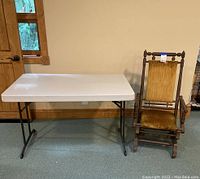 Photo showing the white plastic folding table next to the vintage wooden rocking chair, both fully assembled and standing on carpeted floor.