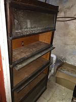Overall view of tall wooden barrister bookcase with four stackable sections, showing missing glass door and water-damaged bottom.