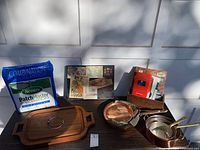 Photo showing all main items on table against white door: copper pots and pans, bag of Scotts PatchMaster lawn seed and fertilizer mix, Goodwood teakwood cheese tray, wooden box with books and frame.