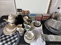 Full overview of the lot on black surface showing pottery teapot, mugs, bowls, check placemats, napkins, napkin rings, decorative tins, and ceramic wall hangings.