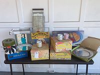 Wide view showing all items on table including boxed serving stand, ceramic tea sets, shag rug rake, stainless steel plates, serving tray, and hot tray warmer
