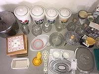Photo showing multiple porcelain canisters with floral designs, various glass containers including oil and vinegar bottles, grinders, and small kitchen utensils organized on a shelf.