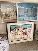 Three wall prints framed, depicting snowy village and town scenes with horse-drawn carriages, parade, and snow-covered buildings. Arranged leaning against a wall.