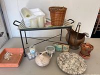 Photo of black metal stand holding a striped ceramic tray with fruit design, copper pot with decorated ceramic handles, terra cotta pots, marble base, battery candles, small framed pictures, a small woven basket, and a ceramic pitcher.