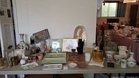 Wide view of various home decor items and perfume on display table including vases, plates, figurines, and baskets.