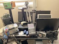 Photo showing multiple black computer monitors, speakers, digital camera, camera tripods, white alarm clock, and Kodak PictureMate photo printer on table.