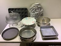 Photo shows various baking tins and cake dishes including glass dome cake dish, floral China cake plate, muffin tray, bunny mold, Bundt pan, pie tin, square tins and springform pans.
