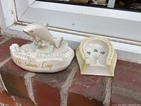 Two Belleek porcelain items placed on brick surface next to a door, horseshoe ashtray showing its top and base with shamrocks, Salmon Leap figurine with fish and waves