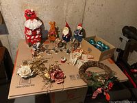 Photo showing the full lot of Christmas decor items including metal figures, wreath, ornaments, floral picks, and ceramic village pieces on cardboard boxes.