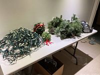 Wide shot showing multiple items including tangled white light strands, garland, mistletoe, and spotlights arranged on a white folding table