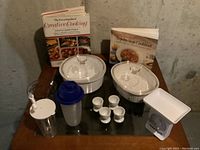 Image showing all items together on a glass tabletop: two Corning Ware dishes with lids, two cookbooks, four plastic egg holders, two plastic drink containers, and a white kitchen scale.