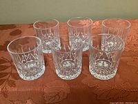 Set of six clear crystal tumblers arranged in two rows on a patterned orange fabric surface showing the cross and olives cut design.
