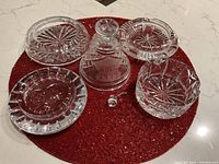 All crystal items displayed together on red placemat including 3 ashtrays, 1 trinket dish, black Noel bell base with detached dinger