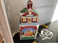 Front view of the porcelain Dickens Winterville Schoolhouse on top of its original box with the full length white power cord coiled beside it.
