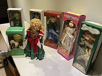 Wide shot of six porcelain dolls, five in original boxes and one on stand without box, displaying variety of brands including Century Collection, Doll Crafters, and Anne of Green Gables.