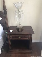 Night stand showing carved wood finish, single drawer with round metallic knob, open lower shelf, placed beside bed with lamp on top.