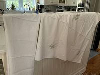 Three white linens folded over a kitchen island: one narrow long towel or runner on the left, a medium tablecloth in the middle, and a large embroidered tablecloth on the right.