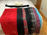 Seven pashmina scarves and two neck warmers laid out on a bed: various solid colors and checkered patterns, including red, black, purple, and turquoise scarves, with a black and cream neck warmer at top.