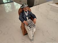 Photo showing entire Royal Doulton figurine seated on wicker armchair holding newspaper.