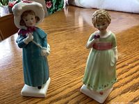 Both Royal Doulton Kate Greenaway figurines Carrie and Tess shown together on a wooden table, highlighting size and color differences.