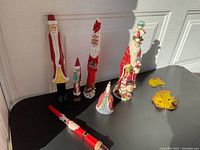 Photo showing seven stick-shaped Santa figurines and a Santa candle, displaying varying sizes and materials on a black surface by a white door.