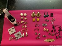 Photo showing 25 pairs of assorted vintage costume earrings in different designs including faux pearls, gold-tone, yellow stones, black stones, and other colored stones arranged on a pink surface with an embellished shoe-shaped jewelry holder.