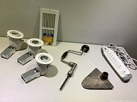 Lot items displayed on white surface: three pot lights with mounting brackets, new return air grill, vintage hand drill, spade drill head, and a power bar.
