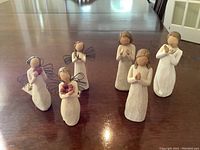 Six Willow Tree angel figurines arranged on a wooden surface, showing three tabletop angels in the back and three winged tree ornaments in front.