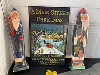 Photo showing two tall wooden Santa figures with original wraps and the Main Street Christmas sign between them.