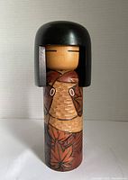 Front view of Kokeshi doll showing carved and hand-painted maple leaf pattern on body, minimal facial features, and black hair.