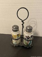 Front view of painted milk glass salt and pepper shakers in silver plated holder showing floral paint and overall condition.