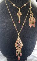 Front view of both necklaces displayed on a black bust showing geometric pendants with reddish enamel and purple glass accents, and matching earrings with similar design.