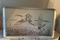 Full view of rectangular aluminum tray with two flying geese engraved over a lake and reeds scene, handles visible at edges.