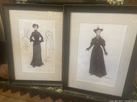 Two framed costume design prints displayed side by side on the floor showing full visuals of both costume illustrations.