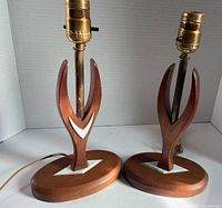 Pair of midcentury modern wooden table lamps side by side showing one lamp with intact white chevron on front and base, the other missing the front chevron. Both have brass bulb sockets with switch. Cord visible.